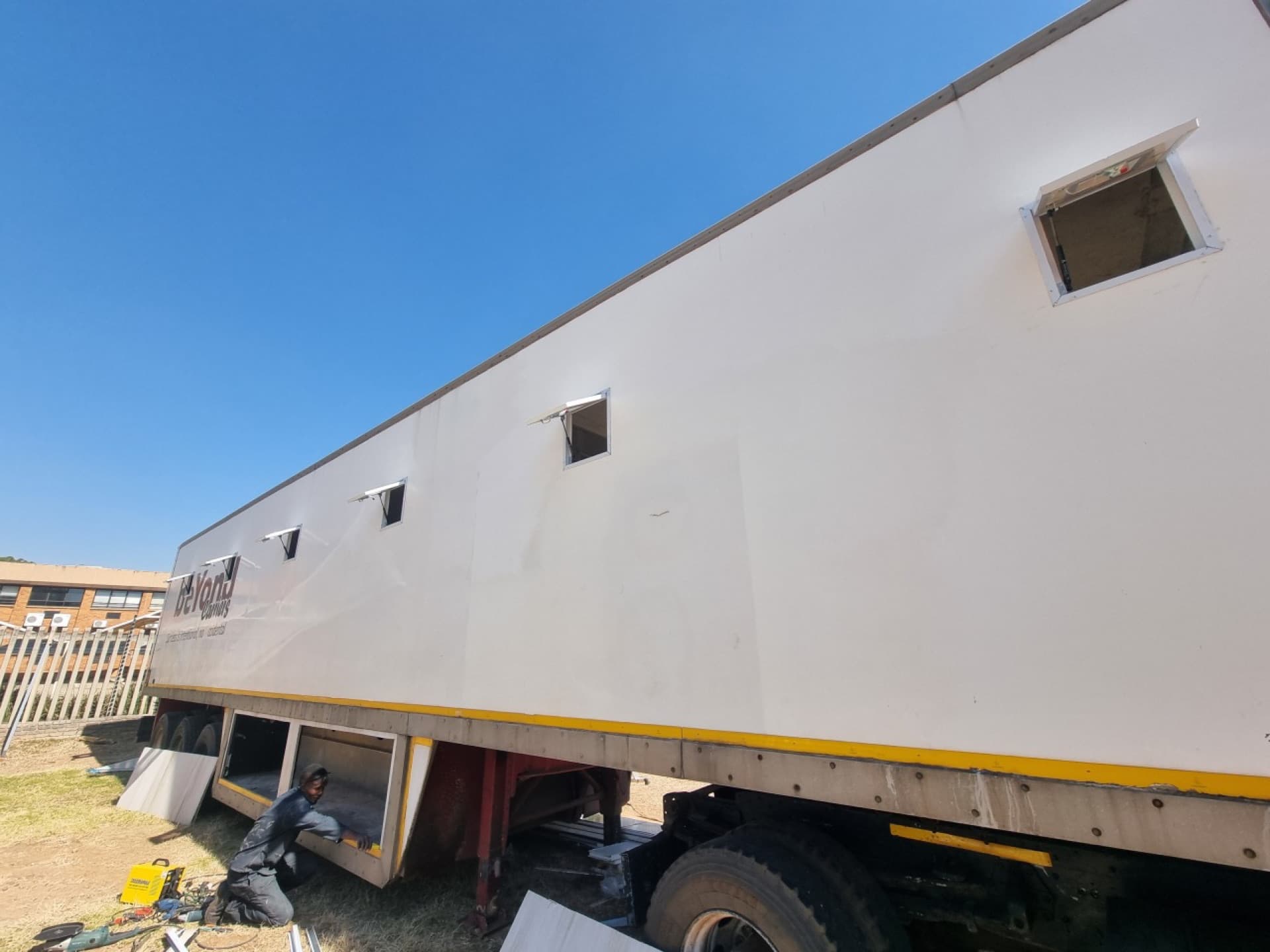A fabricator fitting a lockable underbelly storage locker into the trailer chassis skirt, with awning windows fitted above